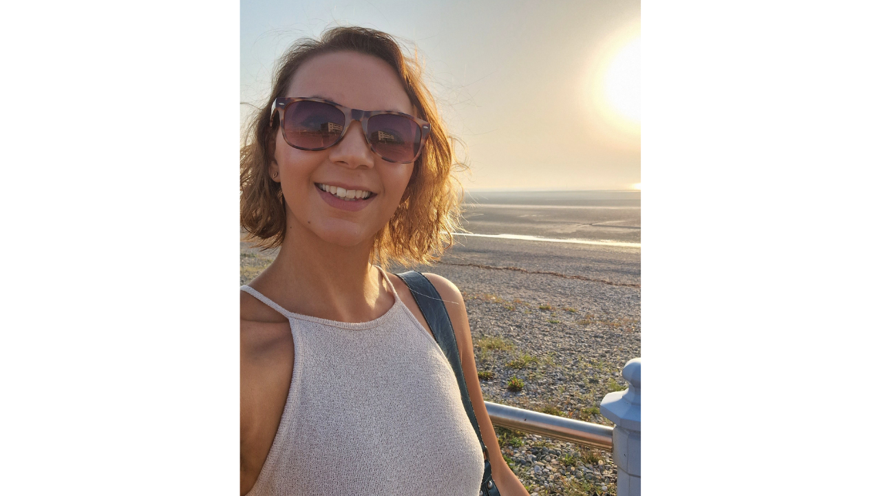 A woman smiling at the camera with a beach at sunset behind her. She's showing how to date yourself